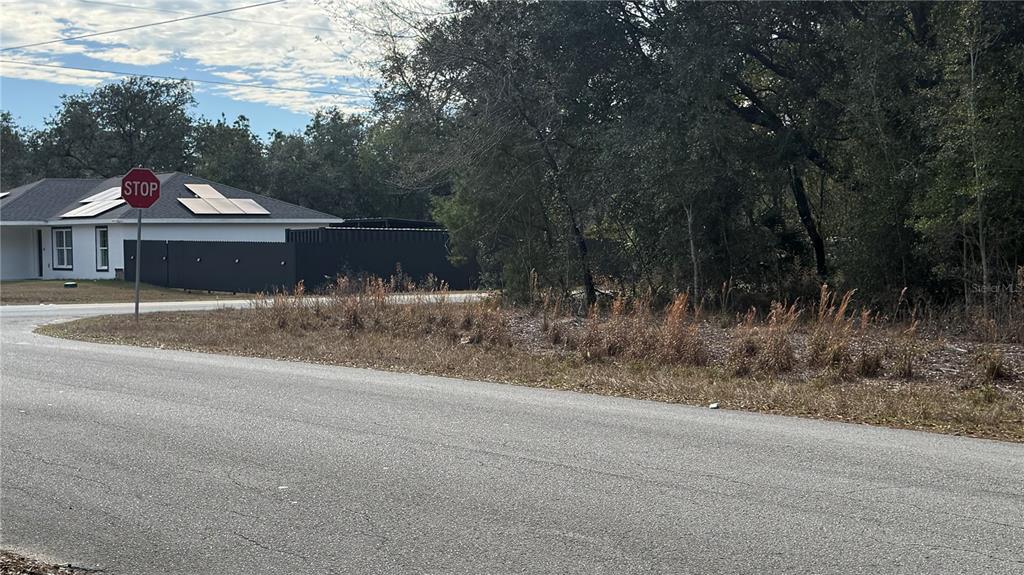 A paved road curves past a vacant, wooded lot next to a house with solar panels — SW BEACH BLVD & SW PORTULACA CT, DUNNELLON, FL