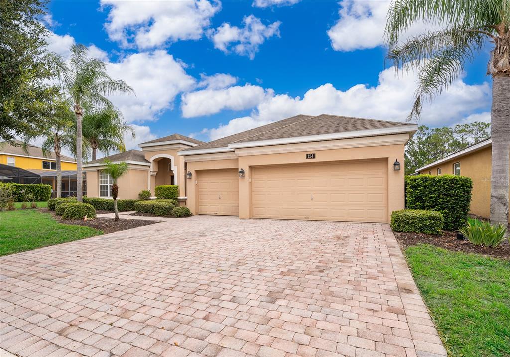 A tan single-story house features a three-car garage and a paved driveway under a blue sky — 124 YELLOW SNAPDRAGON DRIVE, DAVENPORT, FL