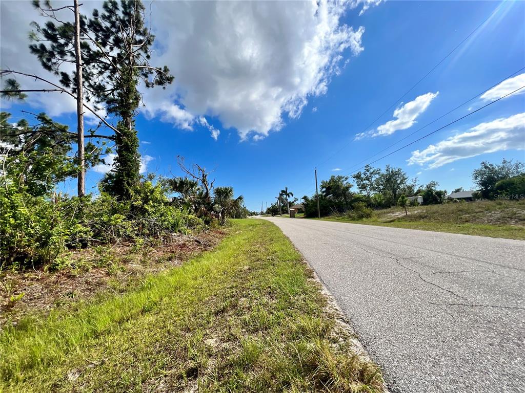 A paved road runs alongside a grassy lot with trees and shrubs under a blue sky — 13463 FROST LANE, PORT CHARLOTTE, FL