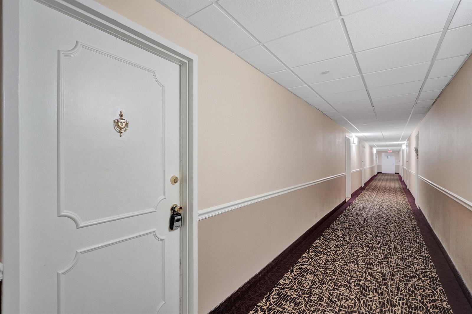 A white door with a lockbox opens into a long hotel-style hallway with patterned carpet — 405 N Ocean Boulevard # 305, Pompano Beach FL 33062, POMPANO BEACH, FL