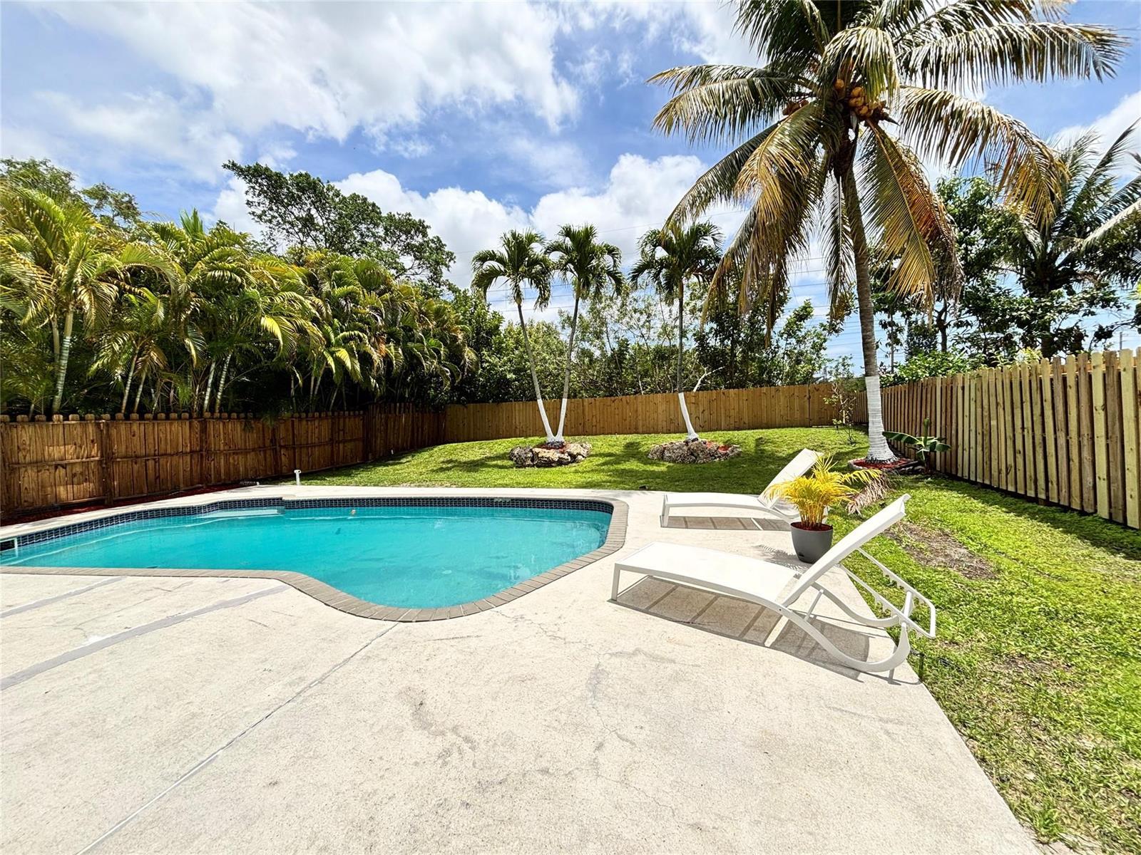 A backyard features a swimming pool, two lounge chairs, and palm trees against a wooden fence — 2990 NW 68th Avenue, Margate FL 33063, MARGATE, FL