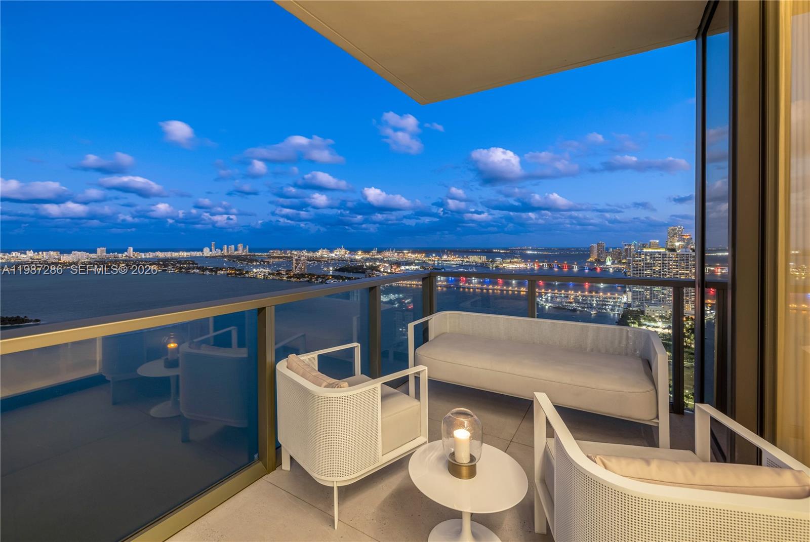 A high-rise balcony features white modern furniture overlooking a city skyline and water at dusk — 788 NE 23rd St # 3801, Miami FL 33137, MIAMI, FL
