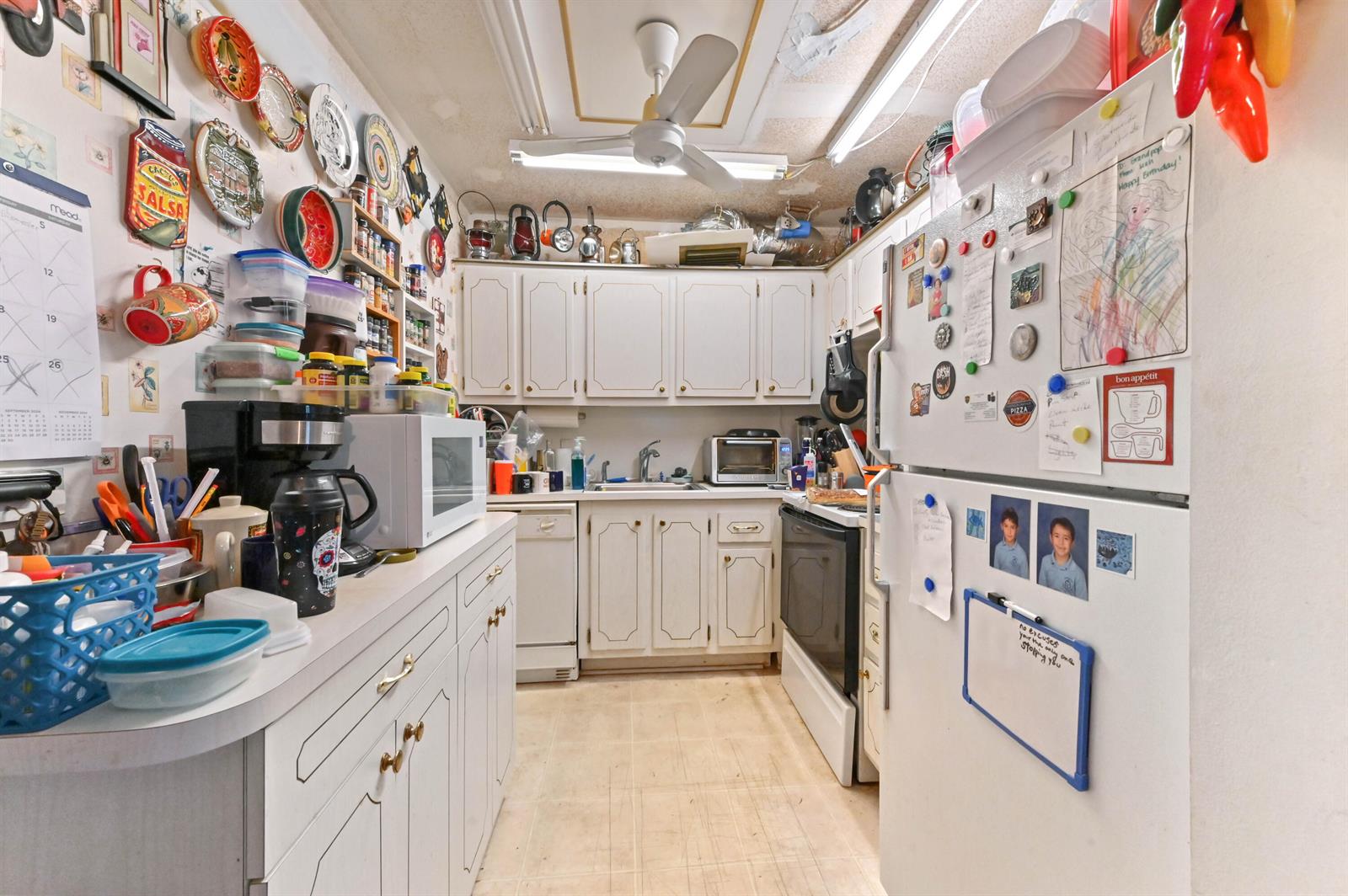 A small, cluttered kitchen features white cabinets, a ceiling fan, and a refrigerator with magnets — 203 Ellesmere D, Deerfield Beach FL 33442, DEERFIELD BEACH, FL