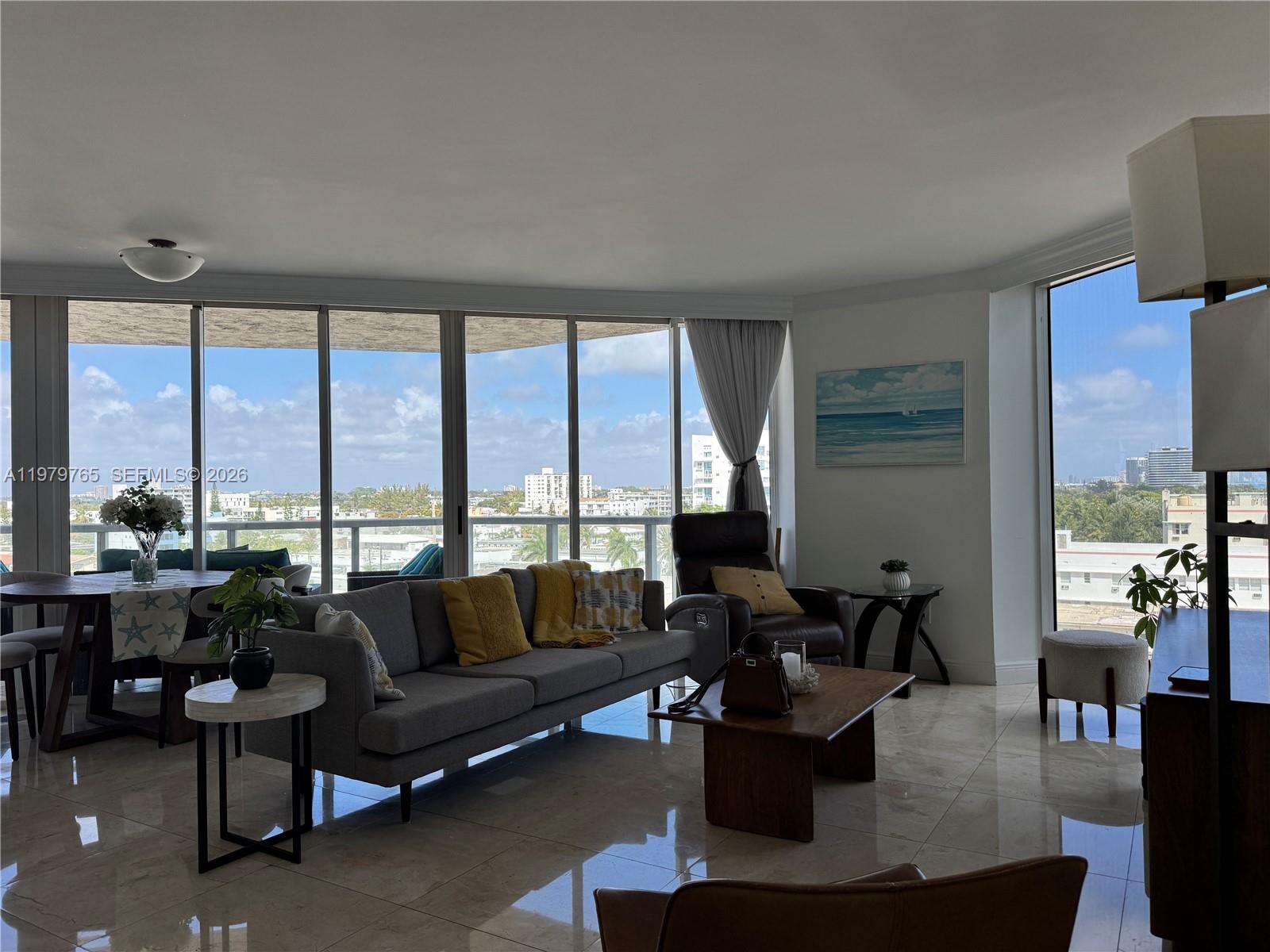 A living room with tile floors, a grey sofa, and floor-to-ceiling windows overlooking a city — 7330 Ocean Ter # D, Miami Beach FL 33141, MIAMI BEACH, FL
