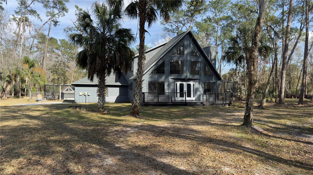 A gray A-frame house with a wooden deck stands in a wooded, grassy yard — 8140 PIMLICO PLACE, WESLEY CHAPEL, FL