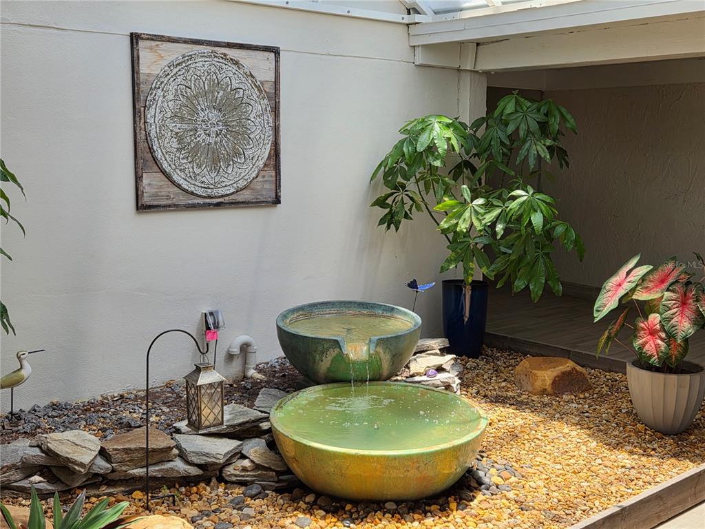A tiered stone fountain sits in a gravel courtyard with potted plants and wall decor — 500 TREASURE ISLAND CAUSEWAY UNIT 307, TREASURE ISLAND, FL