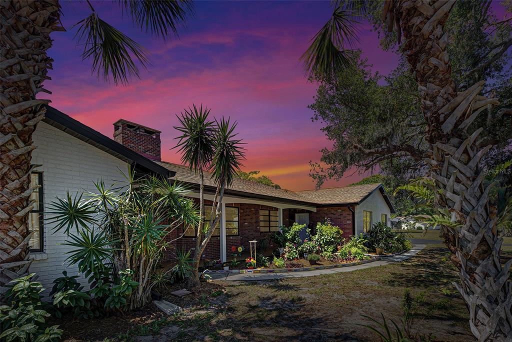 A brick and white-painted ranch-style house exterior features a front garden under a sunset sky — 1061 OWL DRIVE, ENGLEWOOD, FL