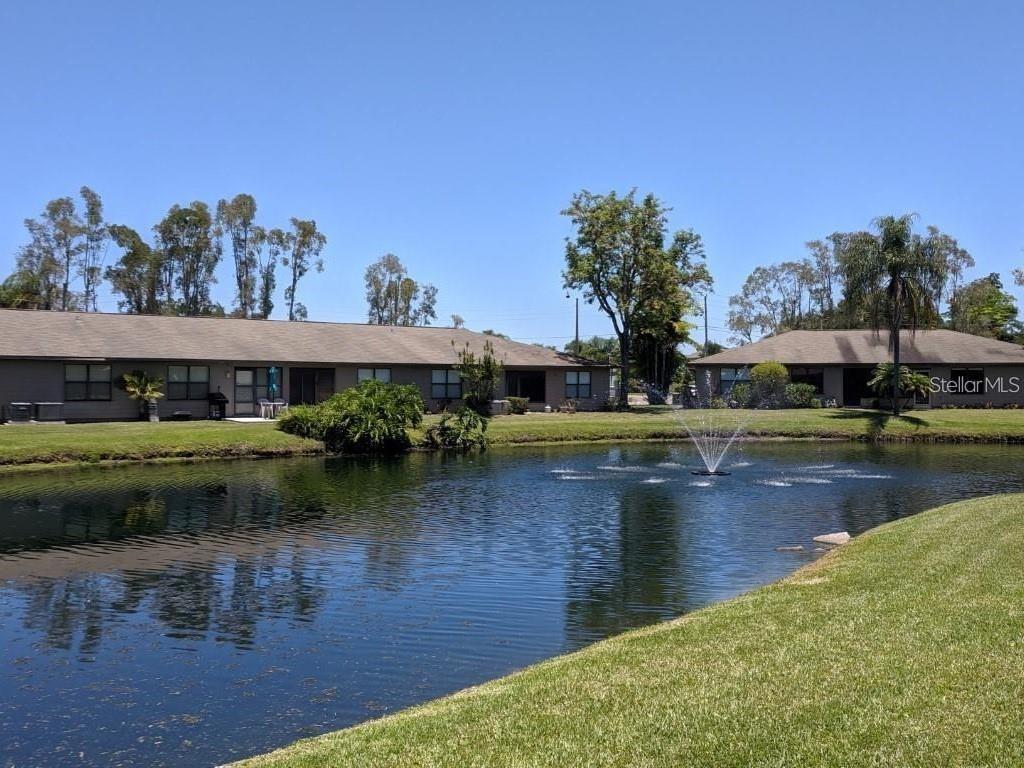 A pond with a fountain sits in front of single-story residential buildings under a sky — 5323 11TH ST CIRCLE E, BRADENTON, FL