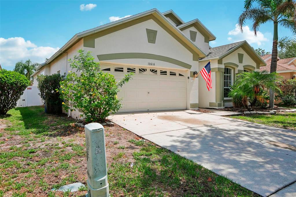 A single-story house with a two-car garage, beige stucco siding, and an American flag — 10616 LITTLE BEND LANE, RIVERVIEW, FL