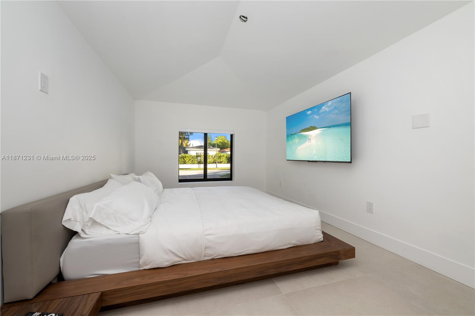 A minimalist bedroom features a low wooden platform bed, white walls, and a wall-mounted television — 900 W 47th Ct, Miami Beach FL 33140, MIAMI BEACH, FL