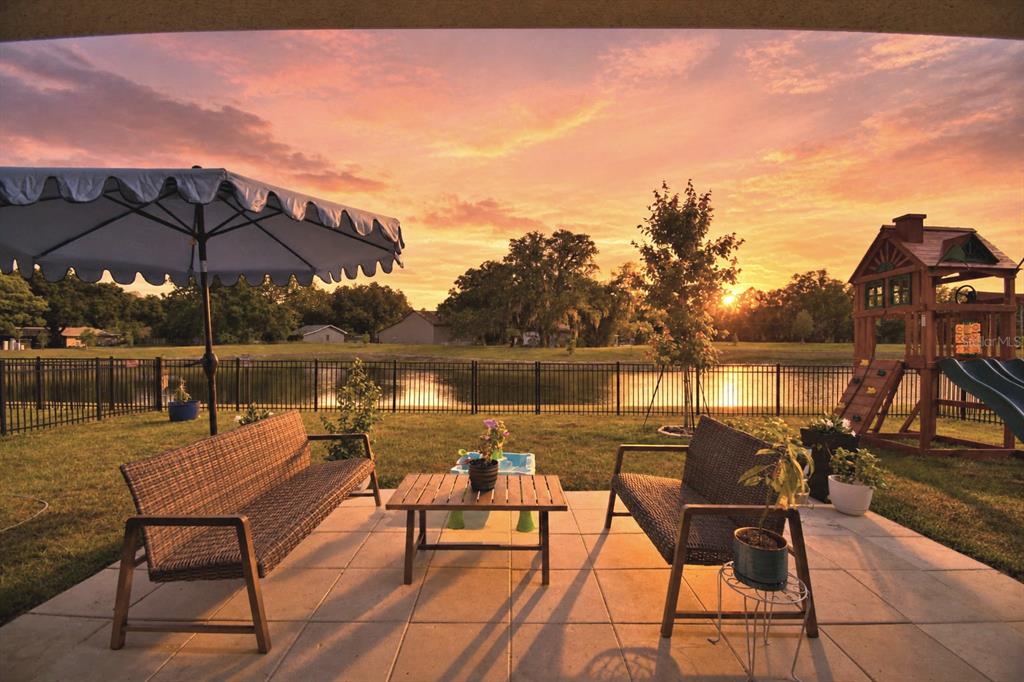A patio with outdoor furniture and a play set overlooks a fenced pond at sunset — 1018 BRIMSTONE CIRCLE, WINTER HAVEN, FL