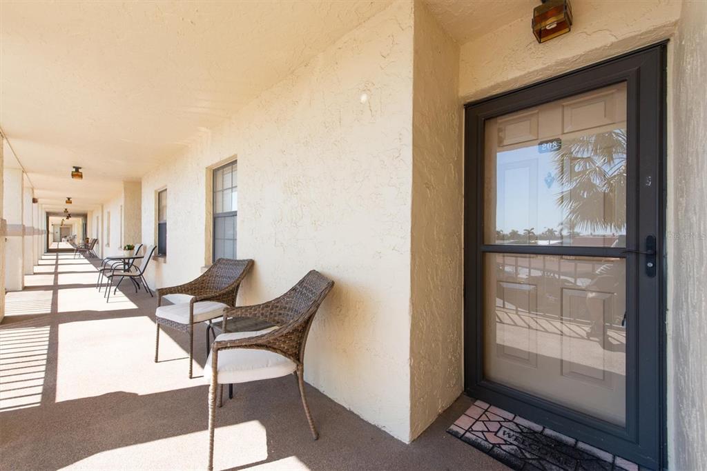 An outdoor apartment walkway features a row of wicker chairs and a glass-paneled entry door — 19029 US HIGHWAY 19 N UNIT 26-208, CLEARWATER, FL