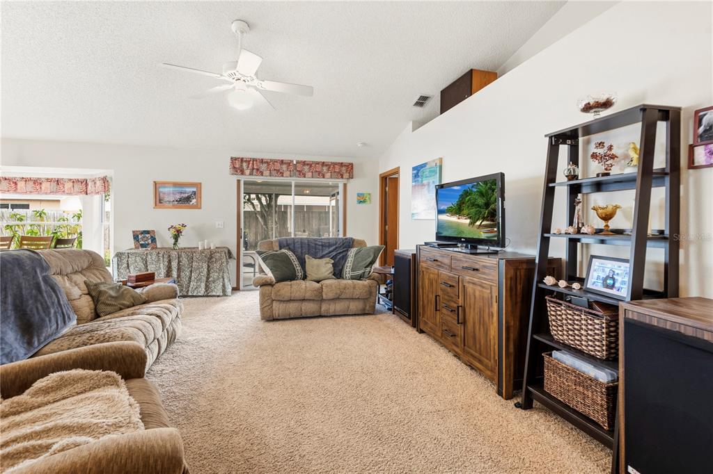 A living room features beige carpet, a ceiling fan, a television console, and a bookshelf — 6937 42ND COURT E, SARASOTA, FL