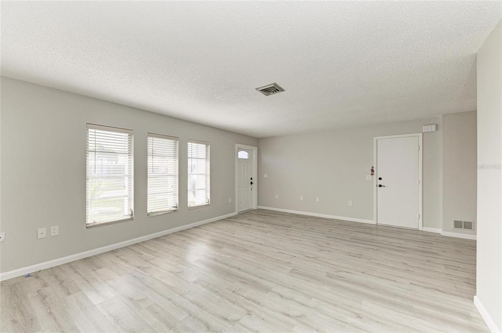 An empty living room features light-colored wood-look flooring, white walls, and three windows with blinds — 140 CIRCLEWOOD DRIVE UNIT C1-4, VENICE, FL