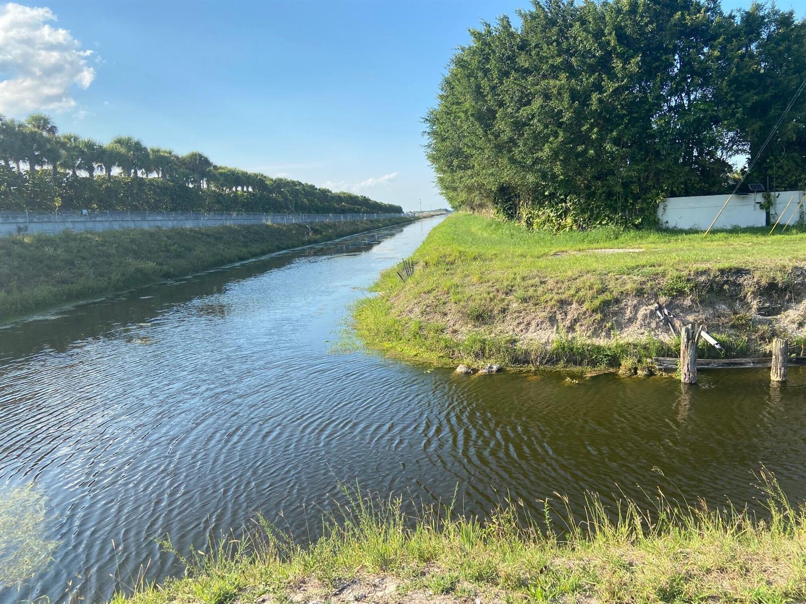 A narrow canal flows between a tree-lined bank and a concrete wall under a blue sky — 4100 Clint Moore Road, Boca Raton FL 33496, BOCA RATON, FL
