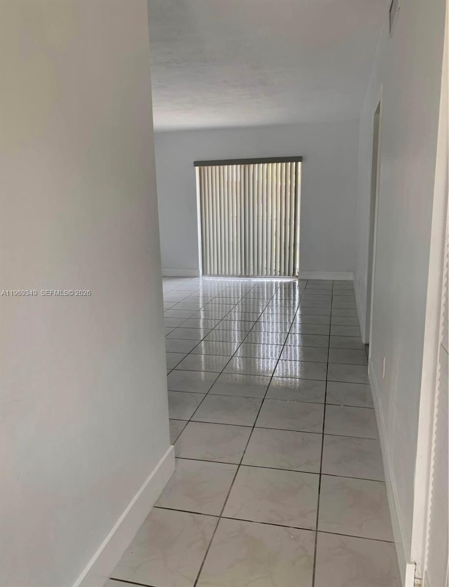 A hallway with light-colored tile flooring leads into a living area with vertical blinds — 7505 SW 82nd St # 306, Miami FL 33143, MIAMI, FL