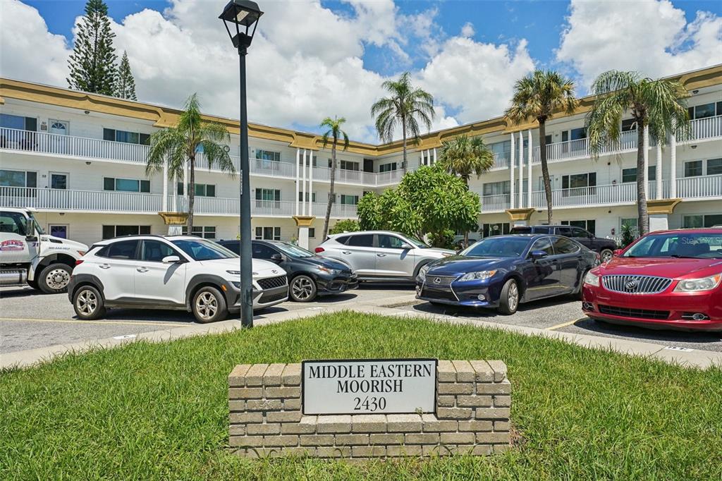 A multi-story apartment building with a parking lot and a brick sign in the foreground — 2430 BRAZILIA DRIVE UNIT 24, CLEARWATER, FL