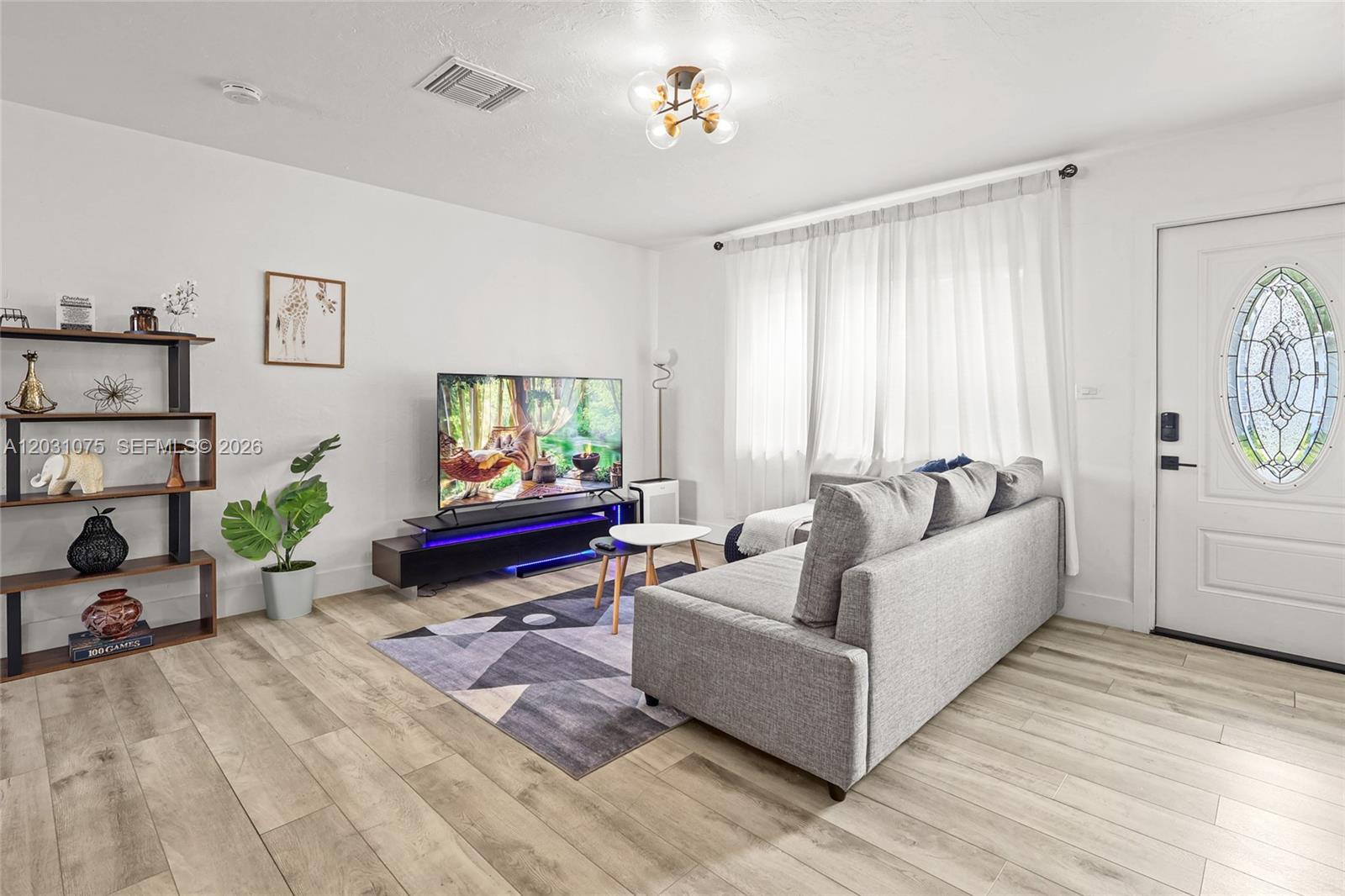 A living room features light wood flooring, a gray sofa, a television, and shelving — 11002 NE 13th Ave, Miami FL 33161, MIAMI, FL