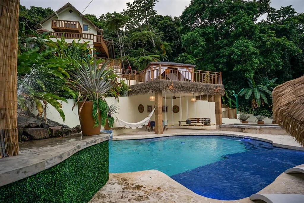 A swimming pool sits in front of a multi-level house with a thatched-roof patio area — CALLE 9 Mameyes II COLINAS DEL YUNQUE UNIT 19 I, RIO GRANDE, PR