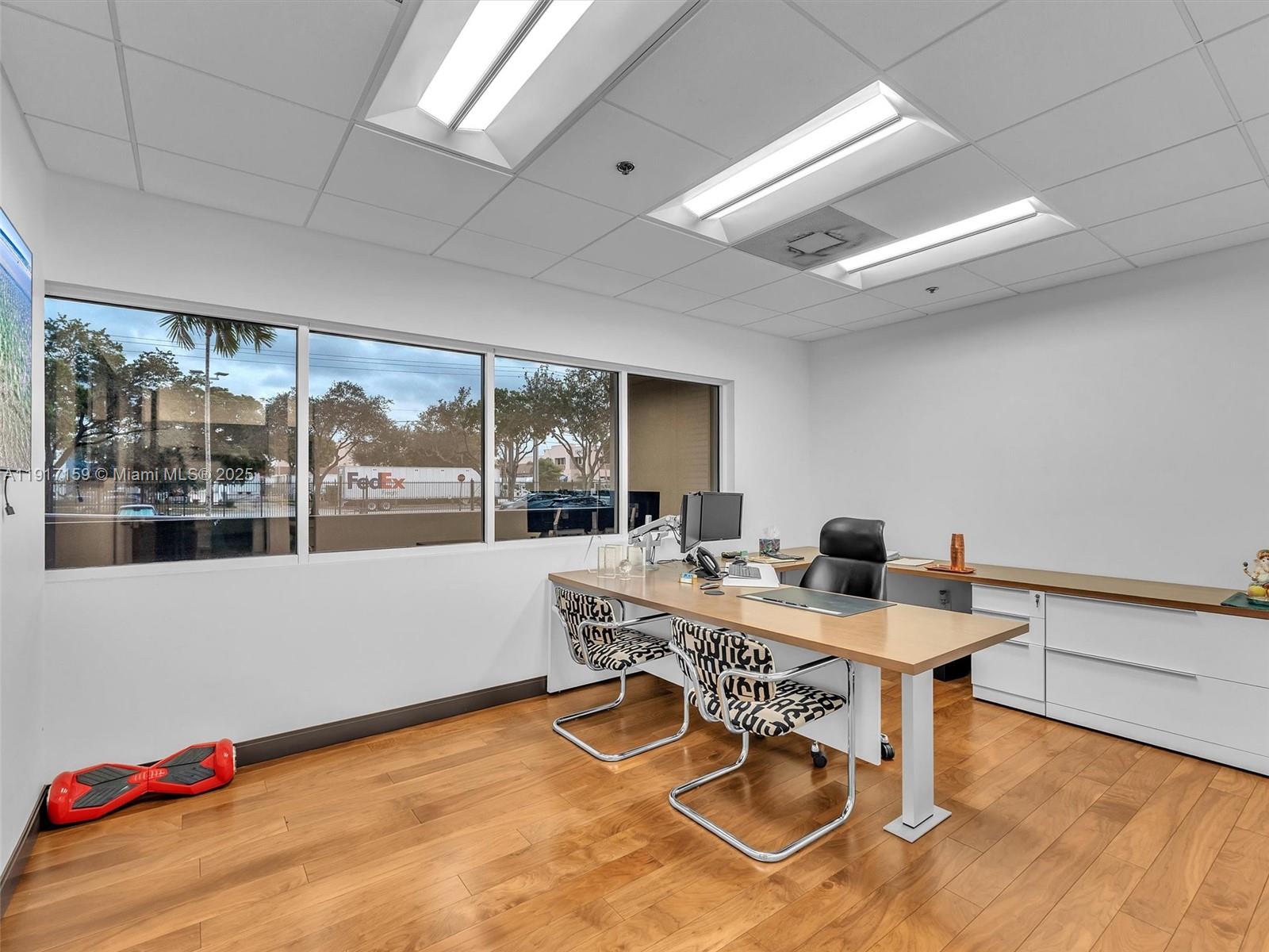 An office with a desk, patterned chairs, wood flooring, and large windows overlooking a parking lot — 10855 NW 27th St, Doral FL 33172, DORAL, FL