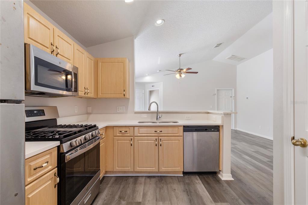 A kitchen with light wood cabinets, stainless steel appliances, and a view into the living area — 7456 OXFORD GARDEN CIRCLE, APOLLO BEACH, FL