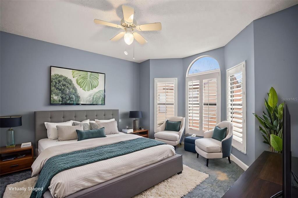 A bedroom features a gray upholstered bed, two armchairs, and large windows with plantation shutters — 1668 HAMILTON COURT UNIT 2061, DUNEDIN, FL
