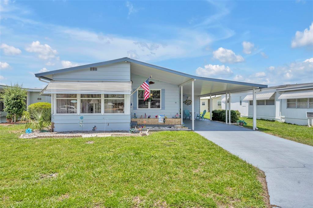A light blue single-story manufactured home features a front porch, carport, and grassy lawn — 91 MAUNA LOA DRIVE, LEESBURG, FL