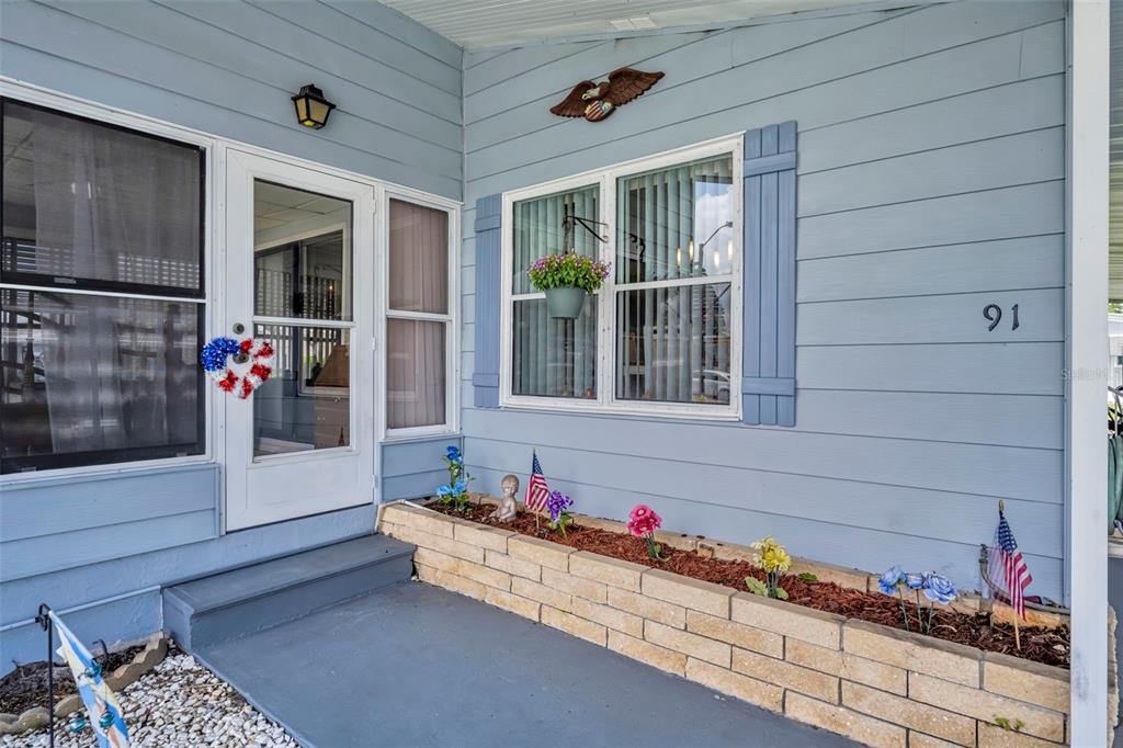 A light blue home exterior features a front porch with a brick planter and flags — 91 MAUNA LOA DRIVE, LEESBURG, FL