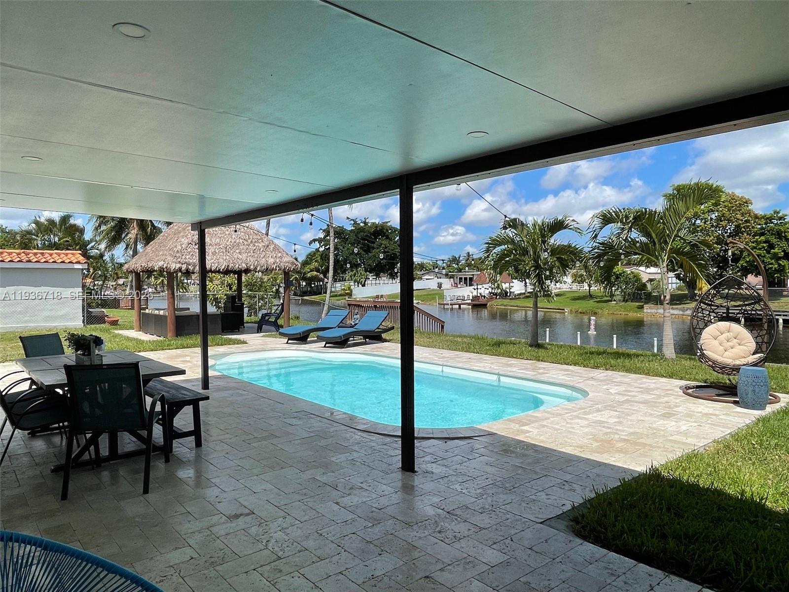 A patio with a pool overlooks a canal with a tiki hut and palm trees — 17831 NW 84th Ct, Miami FL 33015, MIAMI, FL