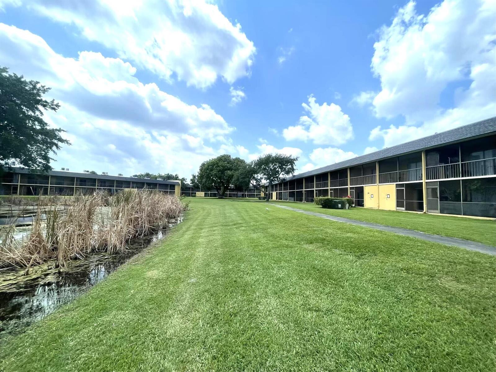 A grassy lawn sits between a pond with reeds and a multi-story apartment building — 1811 Meadows Circle W, Boynton Beach FL 33436, BOYNTON BEACH, FL