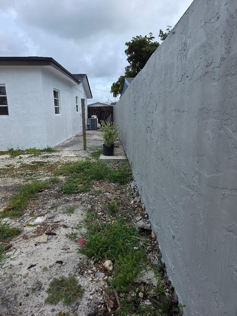 A side view of a white house exterior next to a tall gray concrete wall — HOMESTEAD, FL