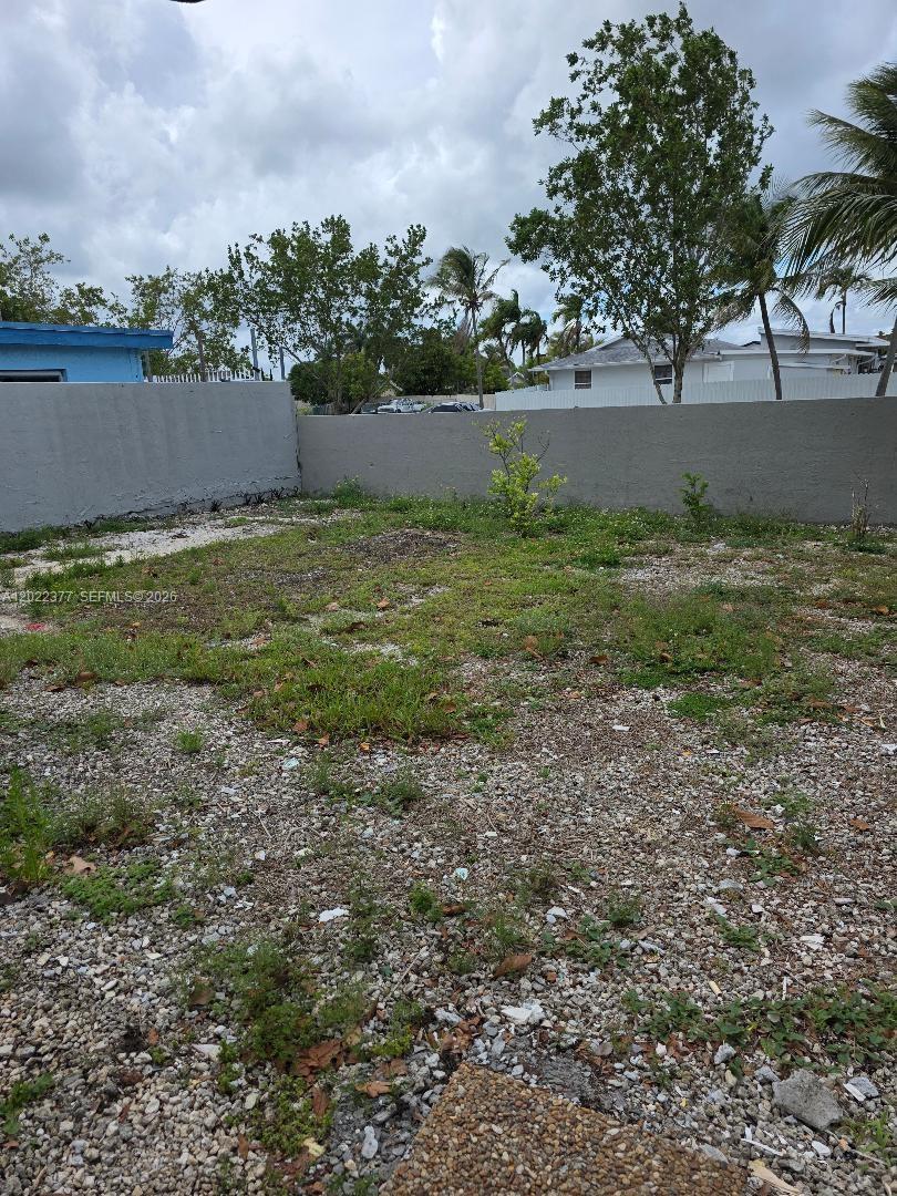 A gravel-covered backyard features sparse patches of grass, a small tree, and a concrete wall — HOMESTEAD, FL