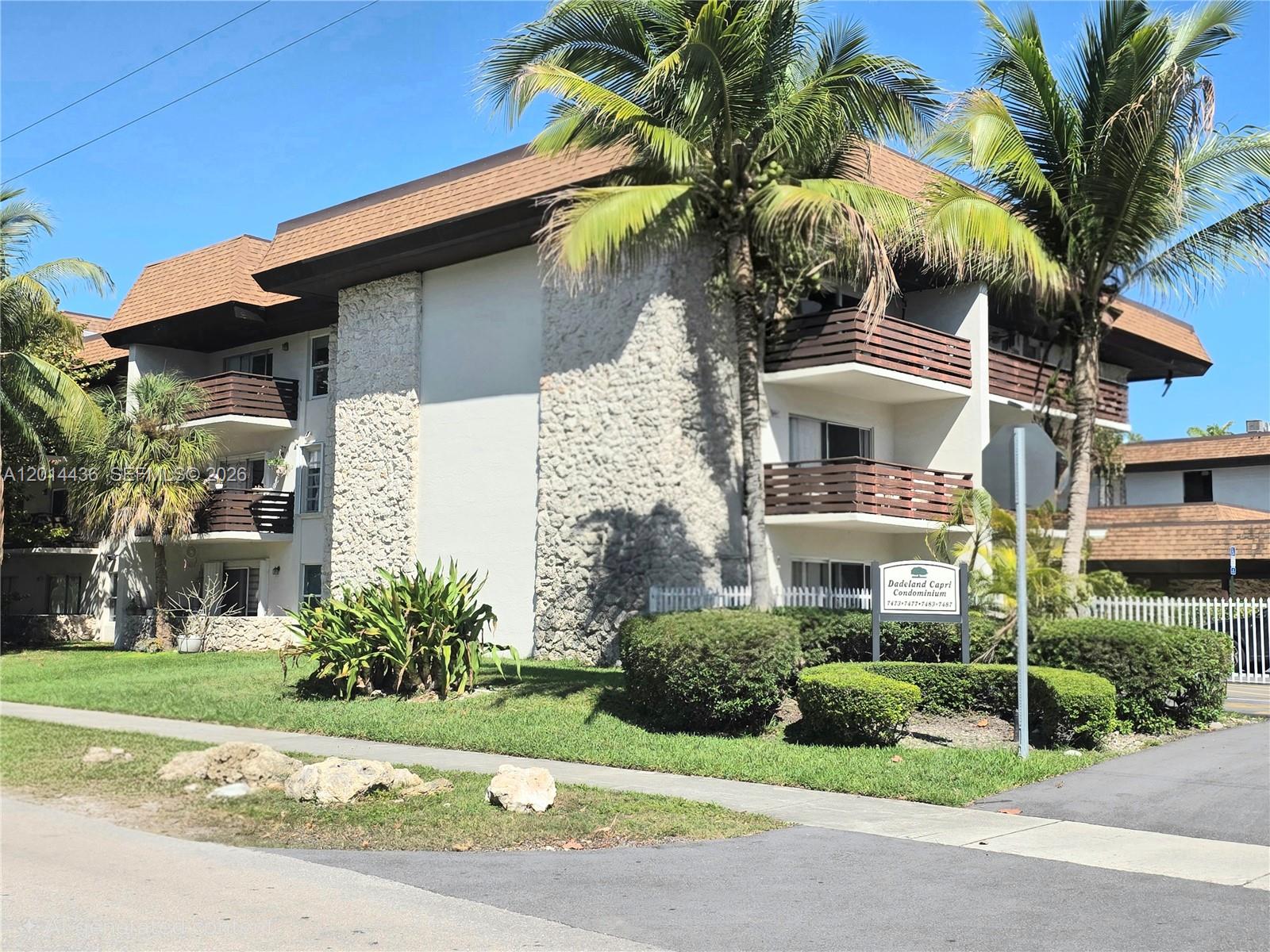 A three-story apartment building features stone accents, brown balconies, and palm trees in the foreground — 7477 SW 82nd St # C115, Miami FL 33143, MIAMI, FL