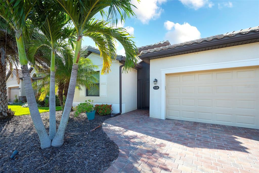 The exterior of a house features a brick paver driveway, a garage, and palm trees — 7130 QUIET CREEK DRIVE, BRADENTON, FL