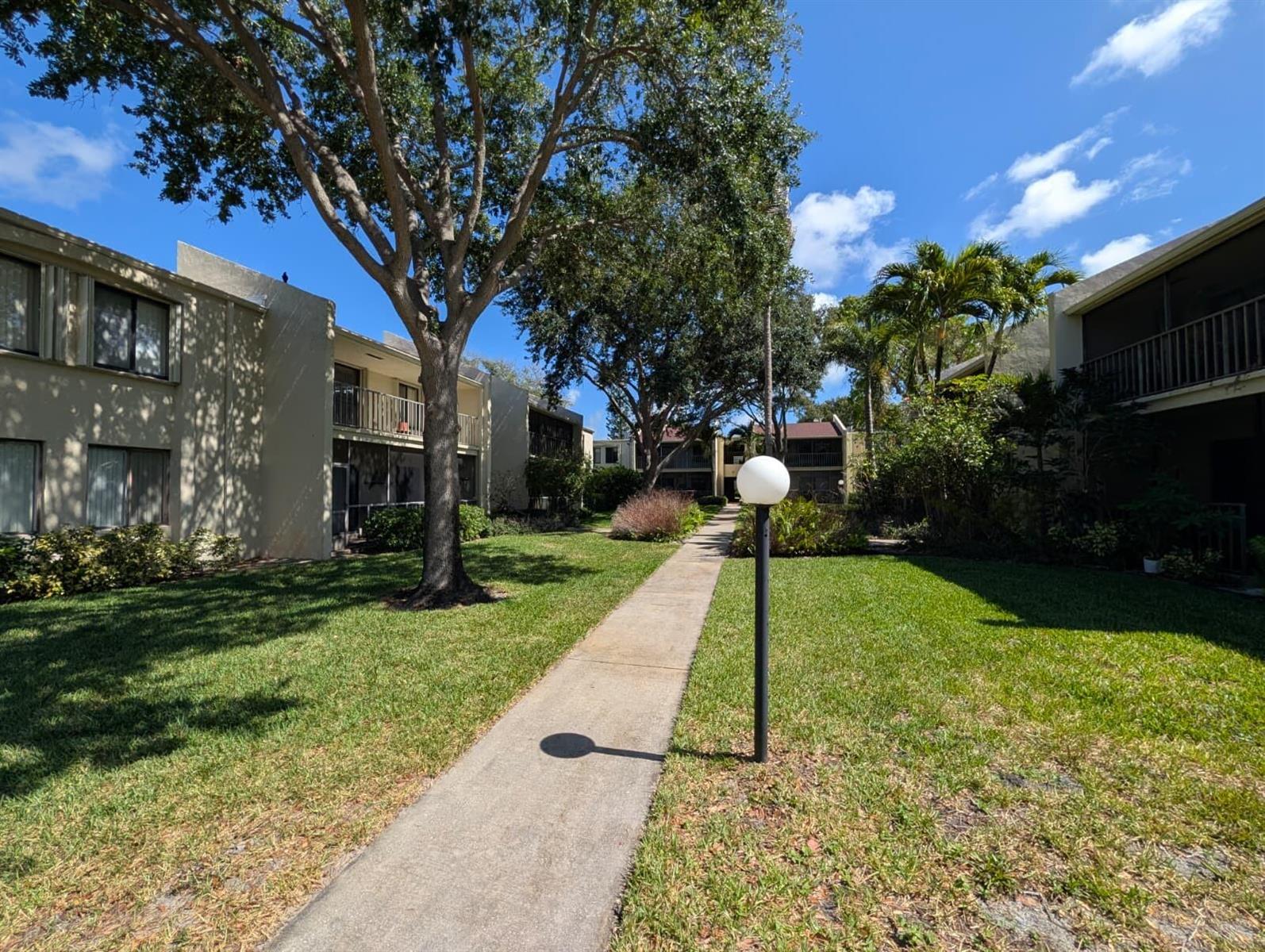 A concrete walkway leads between two apartment buildings with green lawns and large trees — 150 Pineview Road Apt E6, Jupiter FL 33469, JUPITER, FL