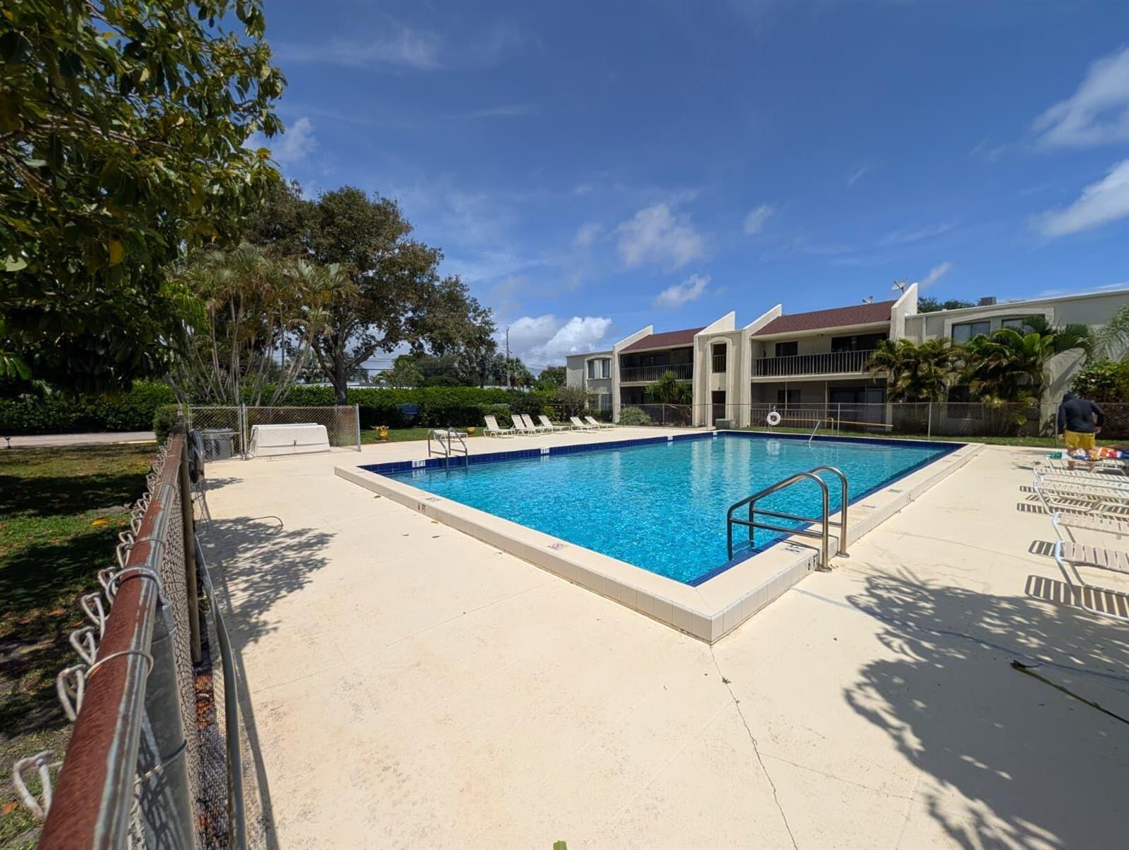 An outdoor swimming pool with a concrete deck is surrounded by a fence and apartment buildings — 150 Pineview Road Apt E6, Jupiter FL 33469, JUPITER, FL