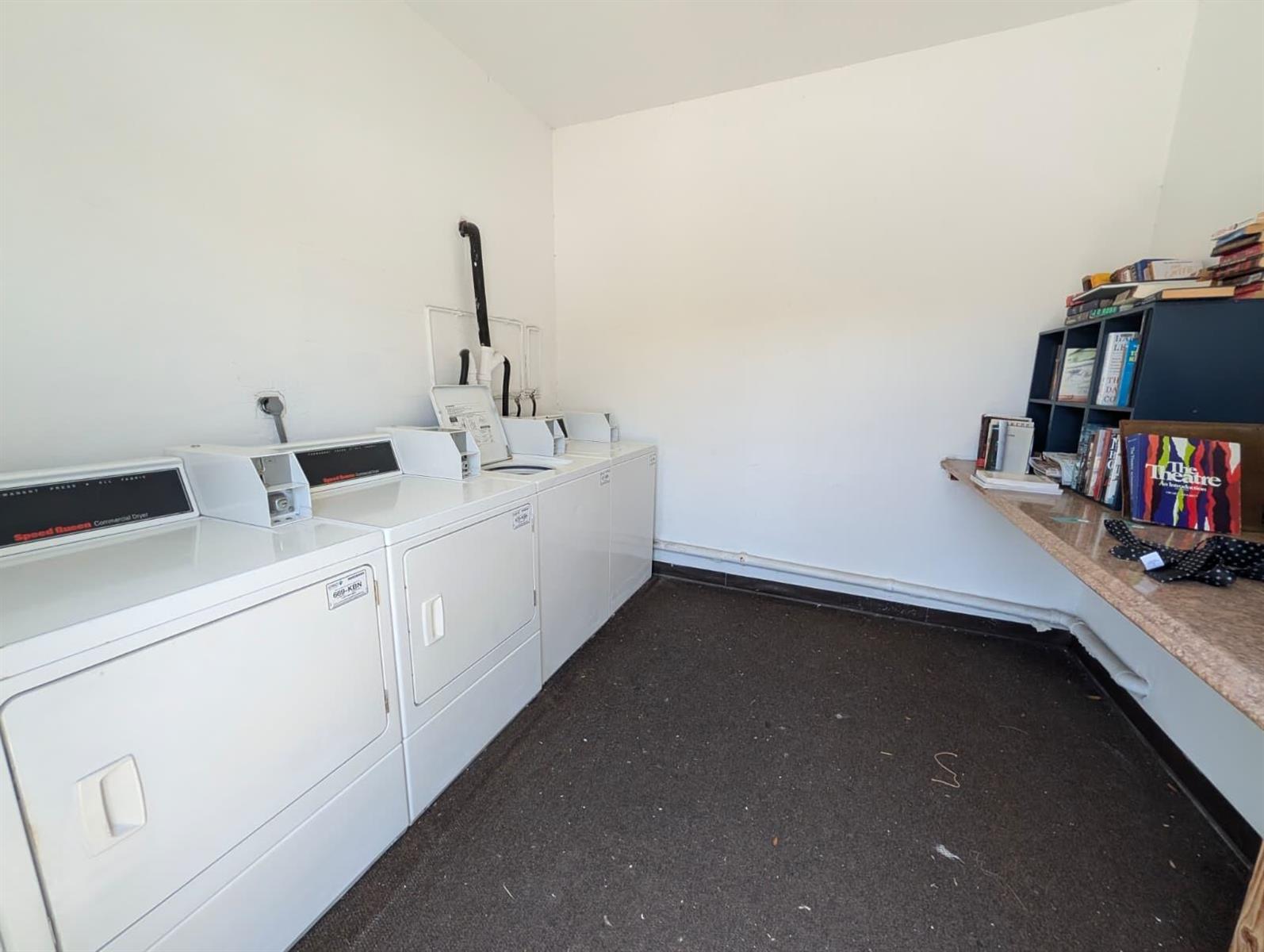 A laundry room features white commercial dryers, a dark carpeted floor, and a built-in bookshelf — 150 Pineview Road Apt E6, Jupiter FL 33469, JUPITER, FL