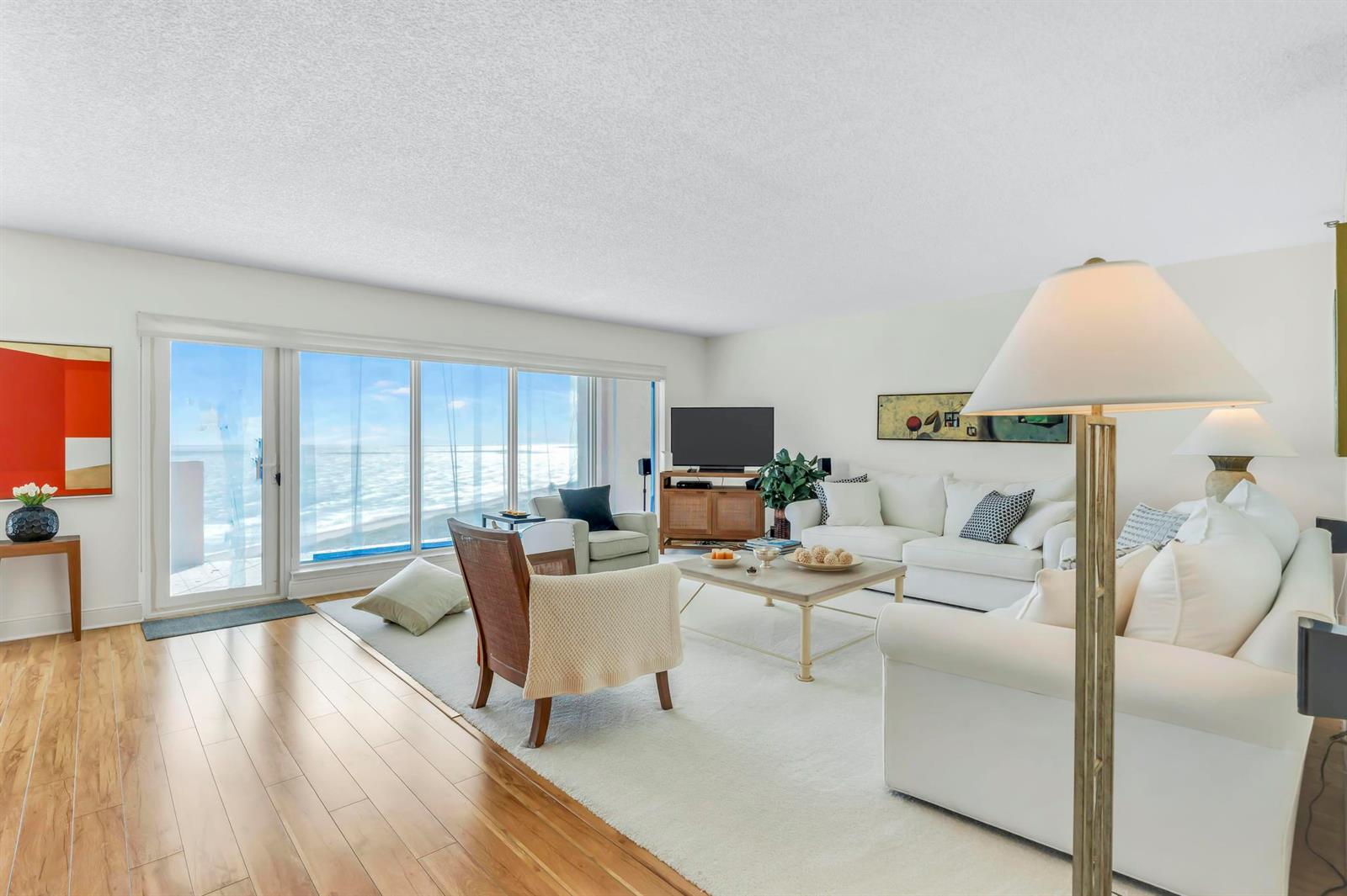 A bright living room features white sofas, a wooden chair, and large windows overlooking the ocean — 4301 N Ocean Boulevard # 1002, Boca Raton FL 33431, BOCA RATON, FL