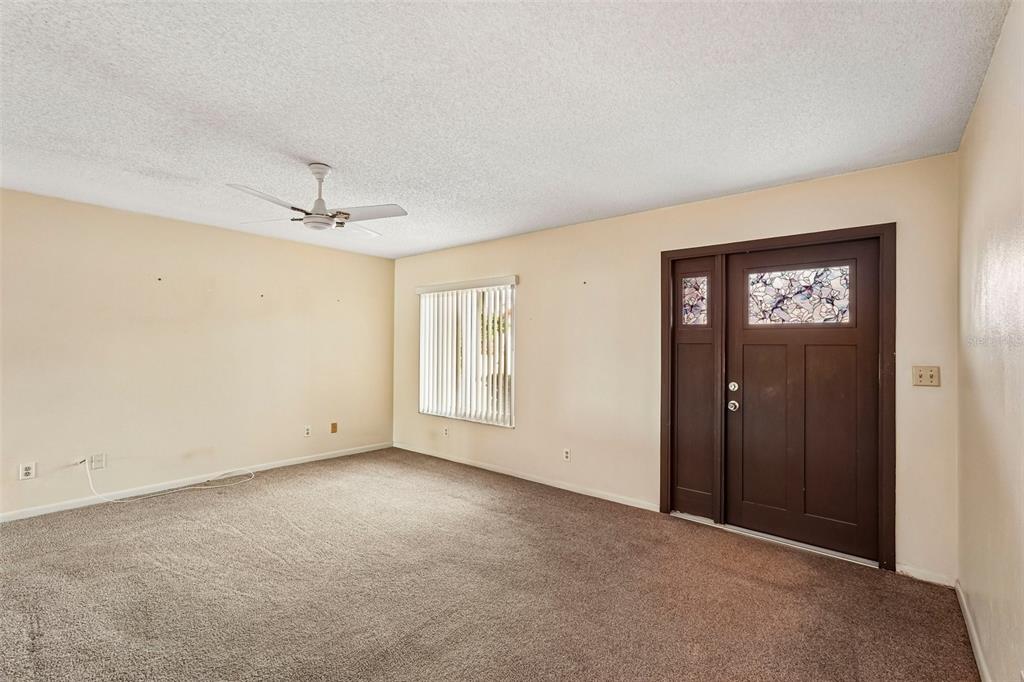 A carpeted living room features a ceiling fan, a window with vertical blinds, and a door — 1329 NEW BEDFORD DRIVE, SUN CITY CENTER, FL