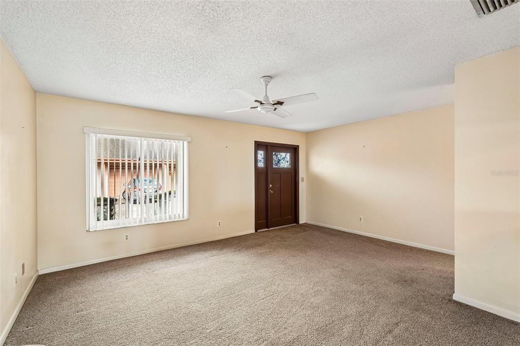 An empty living room with beige walls, brown carpet, a ceiling fan, and a front door — 1329 NEW BEDFORD DRIVE, SUN CITY CENTER, FL