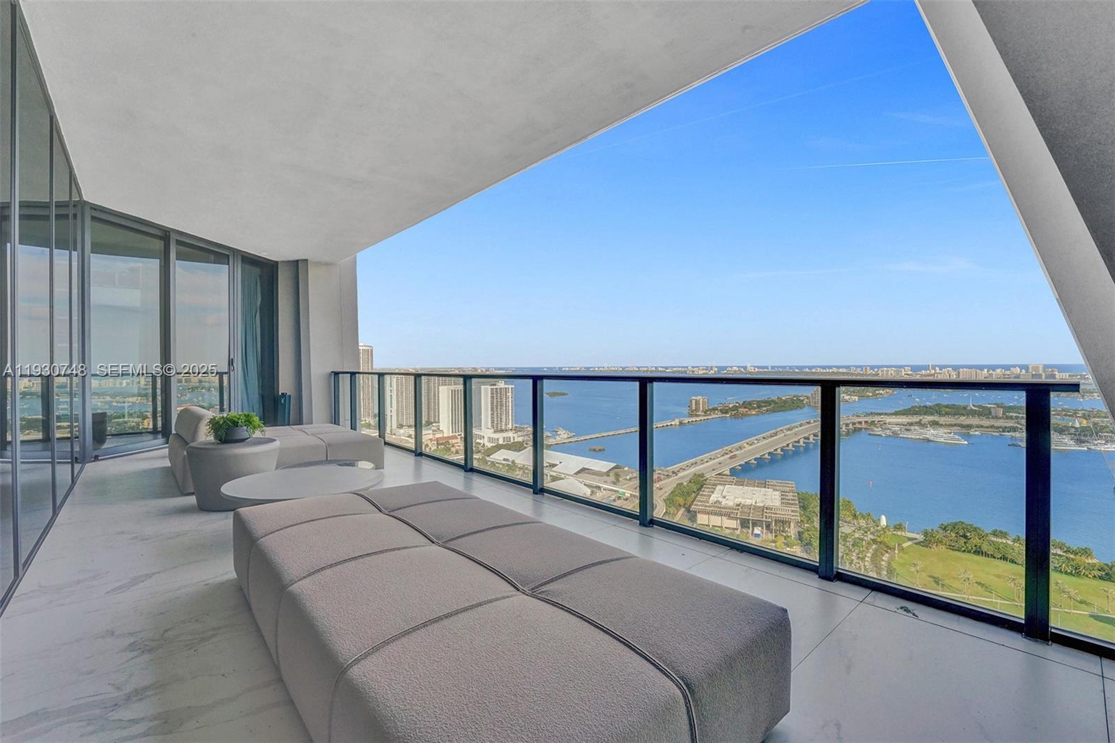 A modern balcony features gray lounge furniture overlooking a bay, a bridge, and city buildings — 1000 Biscayne Blvd # 3902, Miami FL 33132, MIAMI, FL