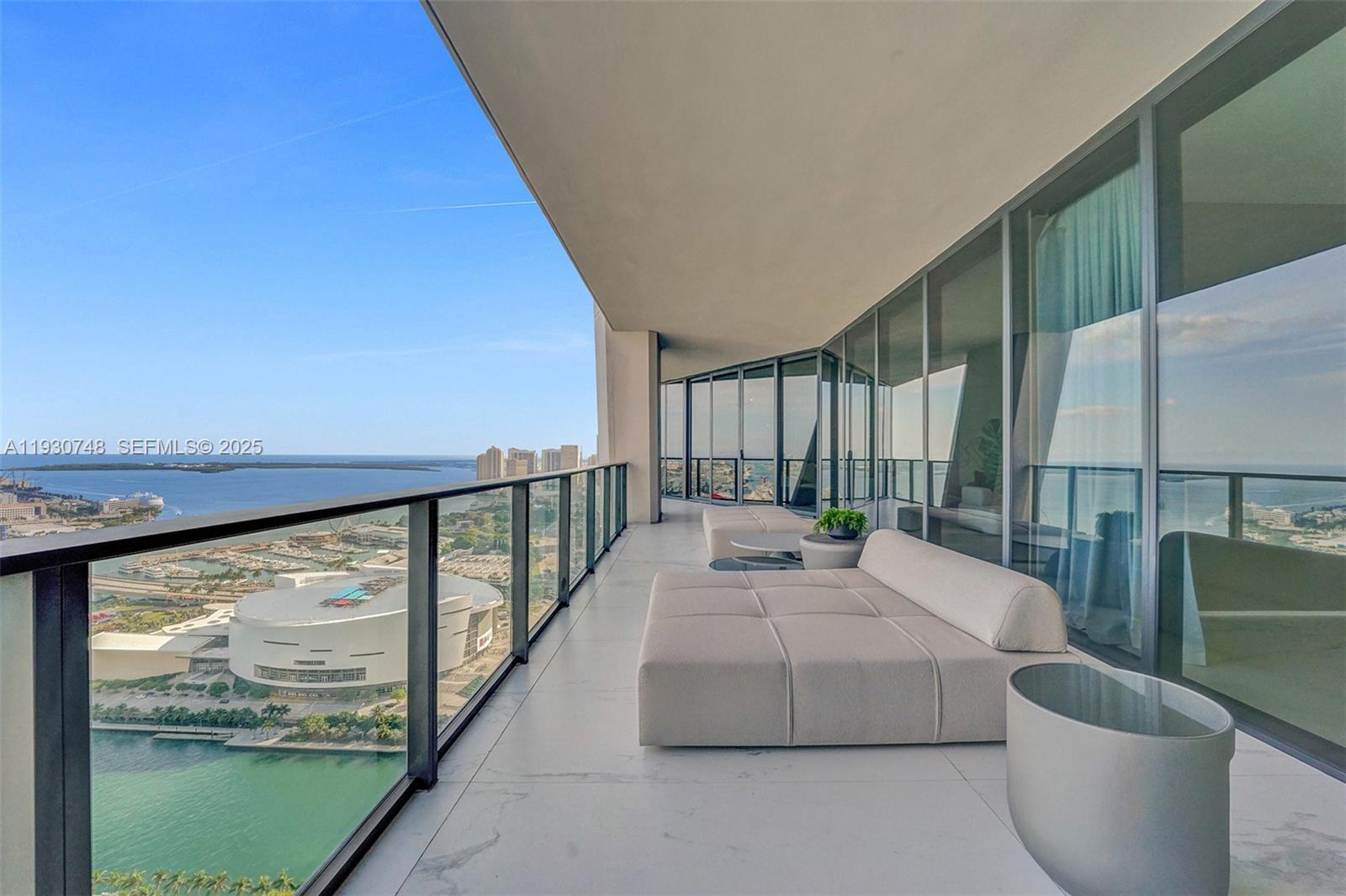 A high-rise balcony features white tile flooring, modern lounge seating, and panoramic waterfront city views — 1000 Biscayne Blvd # 3902, Miami FL 33132, MIAMI, FL