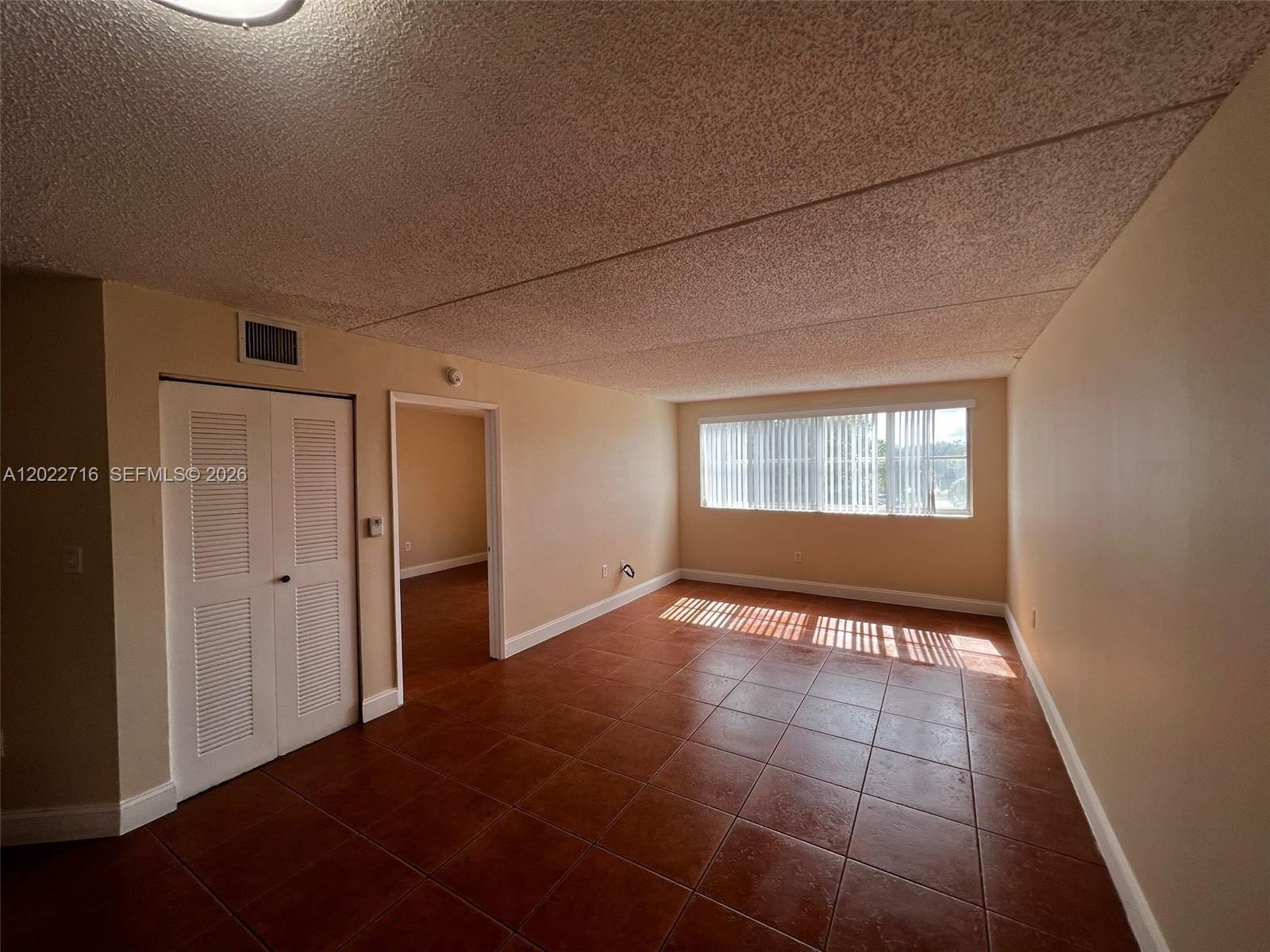 An empty room features terracotta tile flooring, a popcorn ceiling, and a window with blinds — HIALEAH, FL