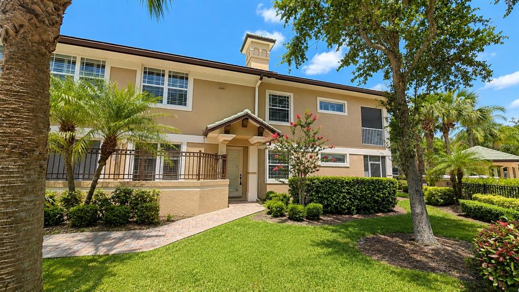 A two-story tan stucco building features a paved walkway, a small porch, and landscaping — 6552 MOORINGS POINT CIRCLE UNIT 102, LAKEWOOD RANCH, FL