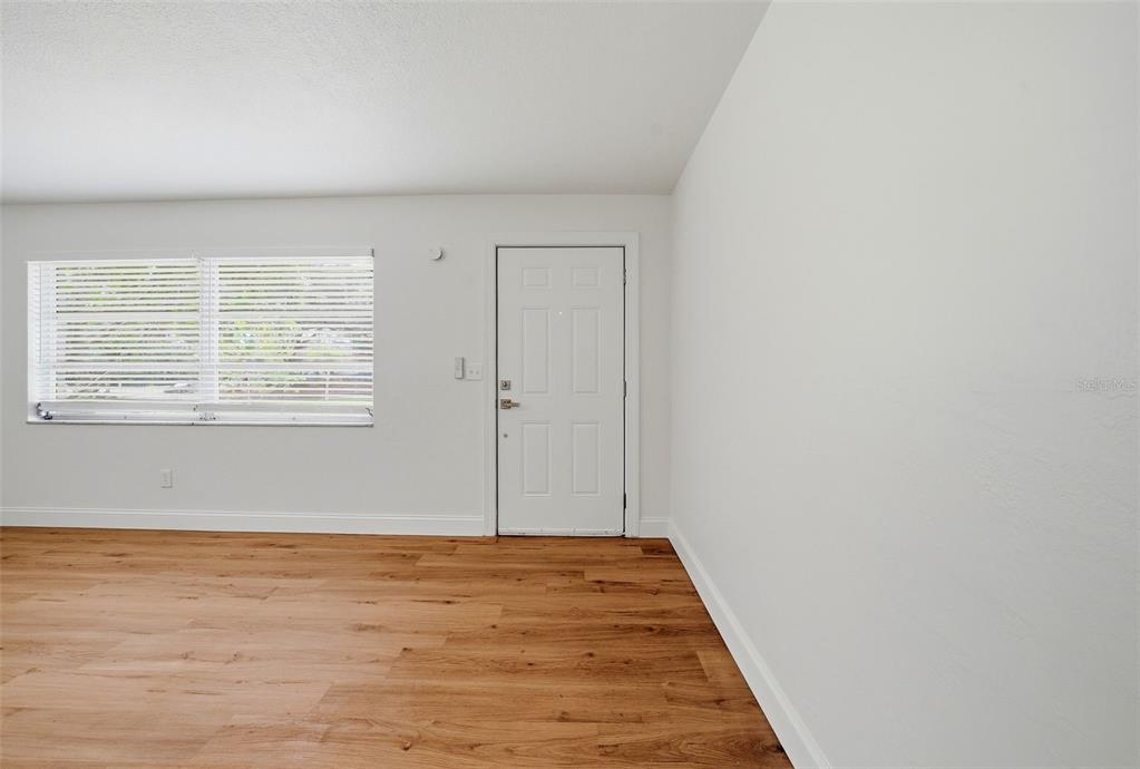 An empty room features light wood-look flooring, white walls, a window with blinds, and a door — 11325 110TH AVENUE, LARGO, FL