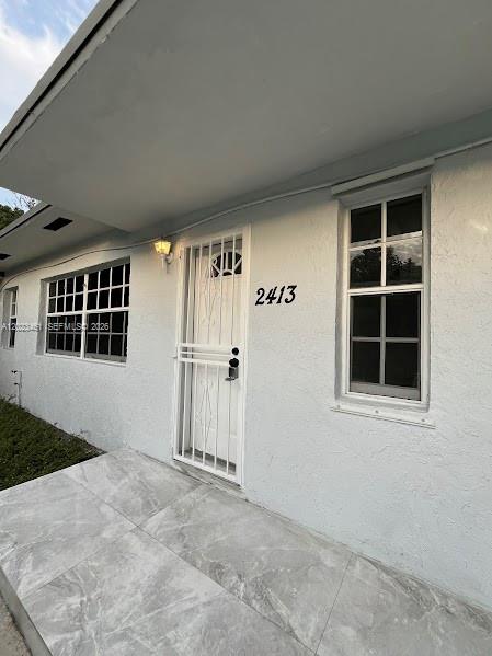 The exterior of a white house features a tiled porch, a security door, and two windows — 2411 NW 65th St, Miami FL 33147, MIAMI, FL