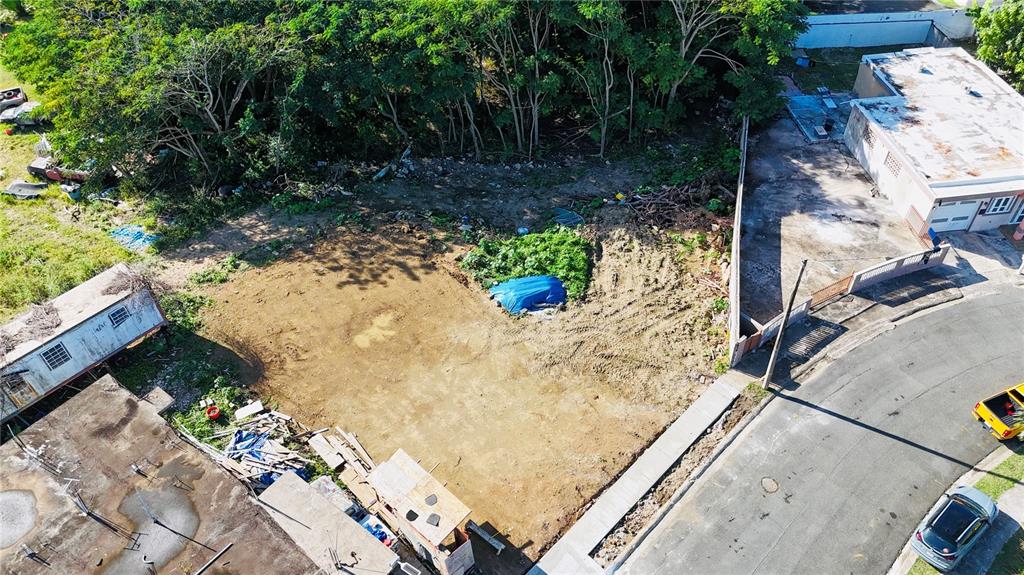 An aerial view shows a vacant dirt lot bordered by trees, a street, and buildings — URB. JARDINES DE LOIZA C-8, LOIZA, PR