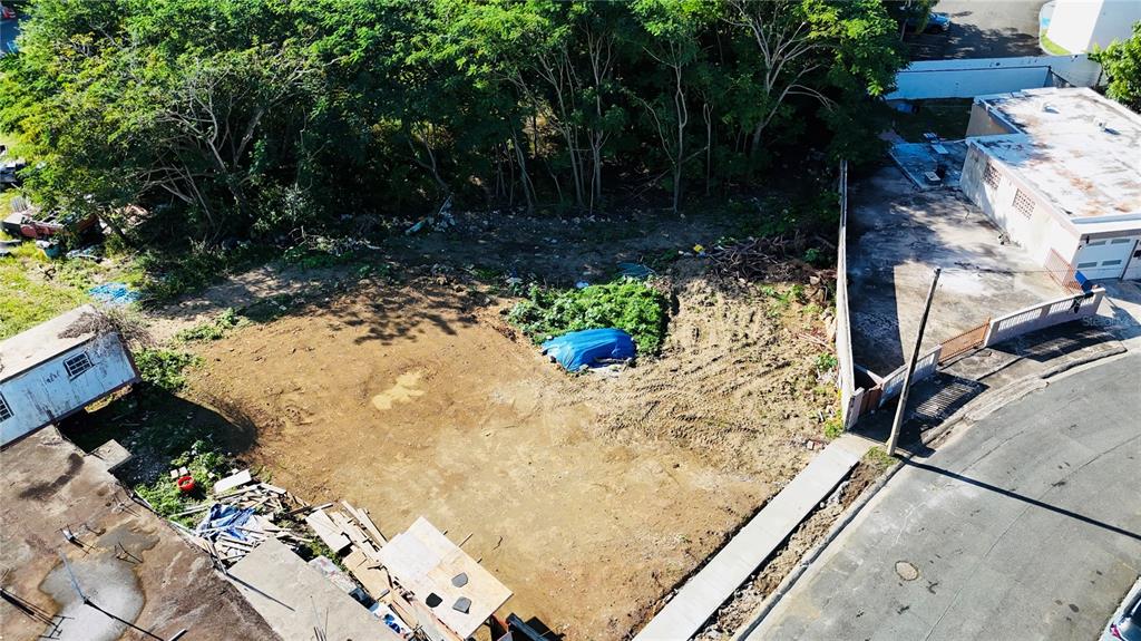 An aerial view shows a vacant dirt lot bordered by trees, a street, and adjacent buildings — URB. JARDINES DE LOIZA C-8, LOIZA, PR