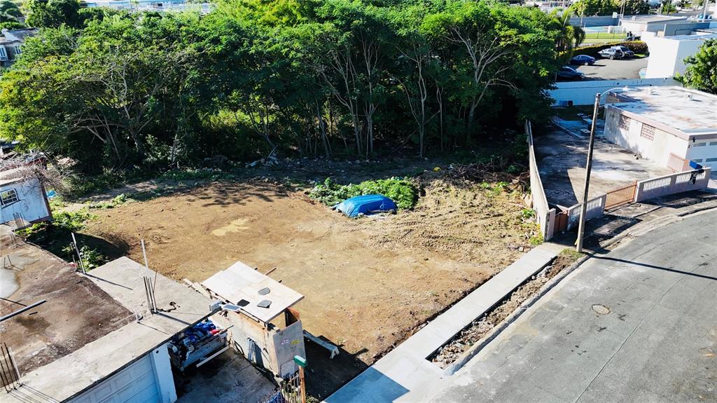 An aerial view shows a vacant, dirt-covered residential lot bordered by trees and neighboring buildings — URB. JARDINES DE LOIZA C-8, LOIZA, PR