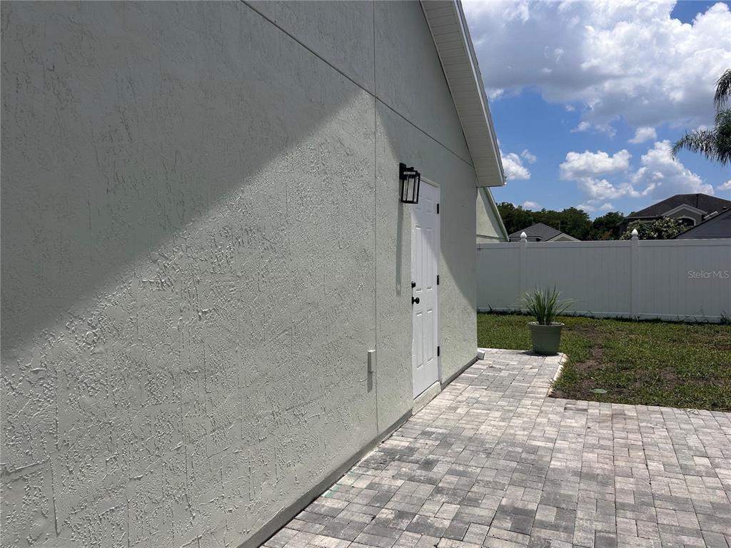 A white stucco exterior wall features a single door, a light fixture, and a paver patio — 6750 BOUGANVILLEA CRESCENT DRIVE, ORLANDO, FL